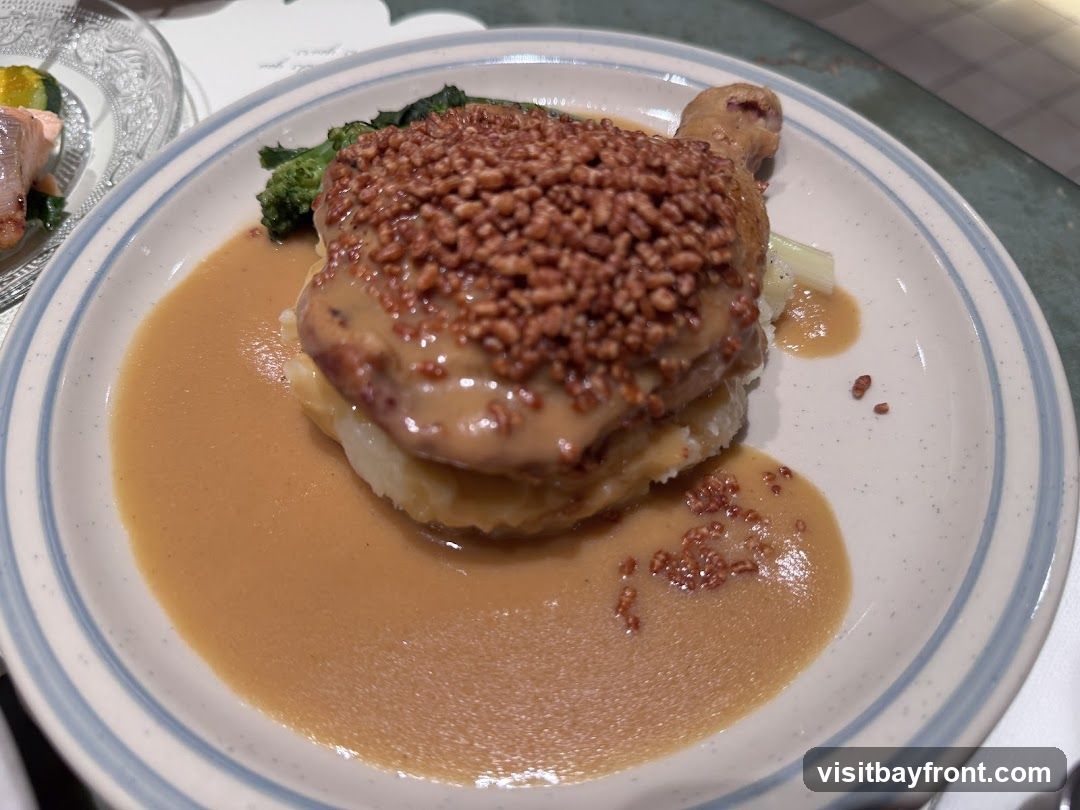 A close-up shot of a plate of food. The dish appears to be a piece of chicken covered in a brown sauce and topped with small, crunchy pieces. It is served on a bed of mashed potatoes with a side of broccoli and what looks like celery. A light brown gravy surrounds the food on the plate. In the background, another plate with salmon and zucchini is partially visible.