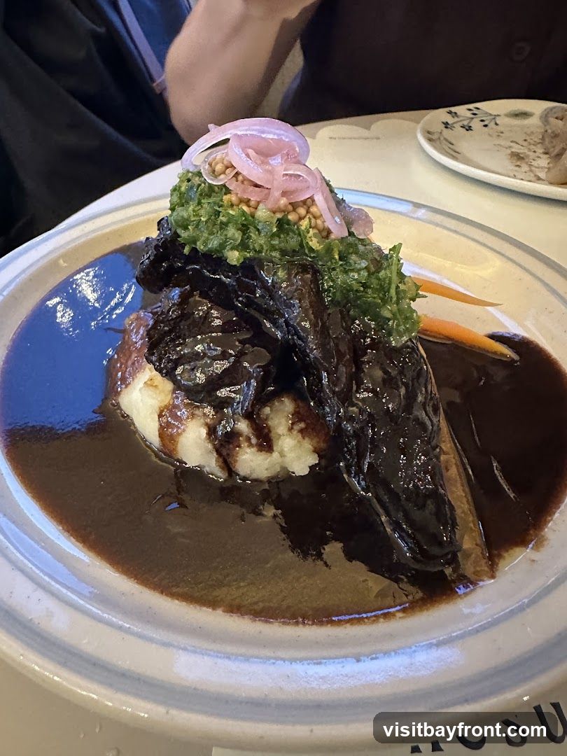 A close-up of a braised dish served with mashed potatoes and a dark sauce. The dish is topped with a green relish, pickled red onions, and seeds. A slice of carrot is visible on the side. The meal is presented on a light-colored plate with a textured rim.