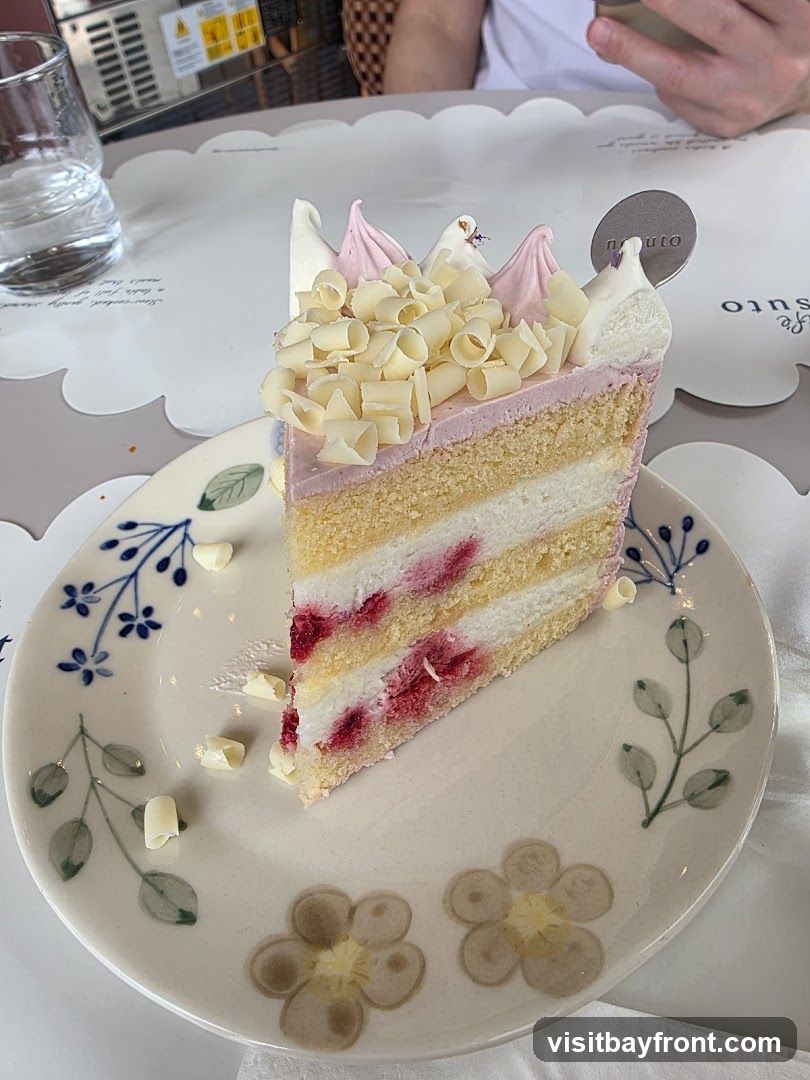 A slice of layered cake with pink frosting, white chocolate shavings, and visible raspberries. The cake is served on a decorative plate with floral motifs. A glass of water and a person's hands are visible in the background. A small circular logo with the word "nauto" is on the cake.