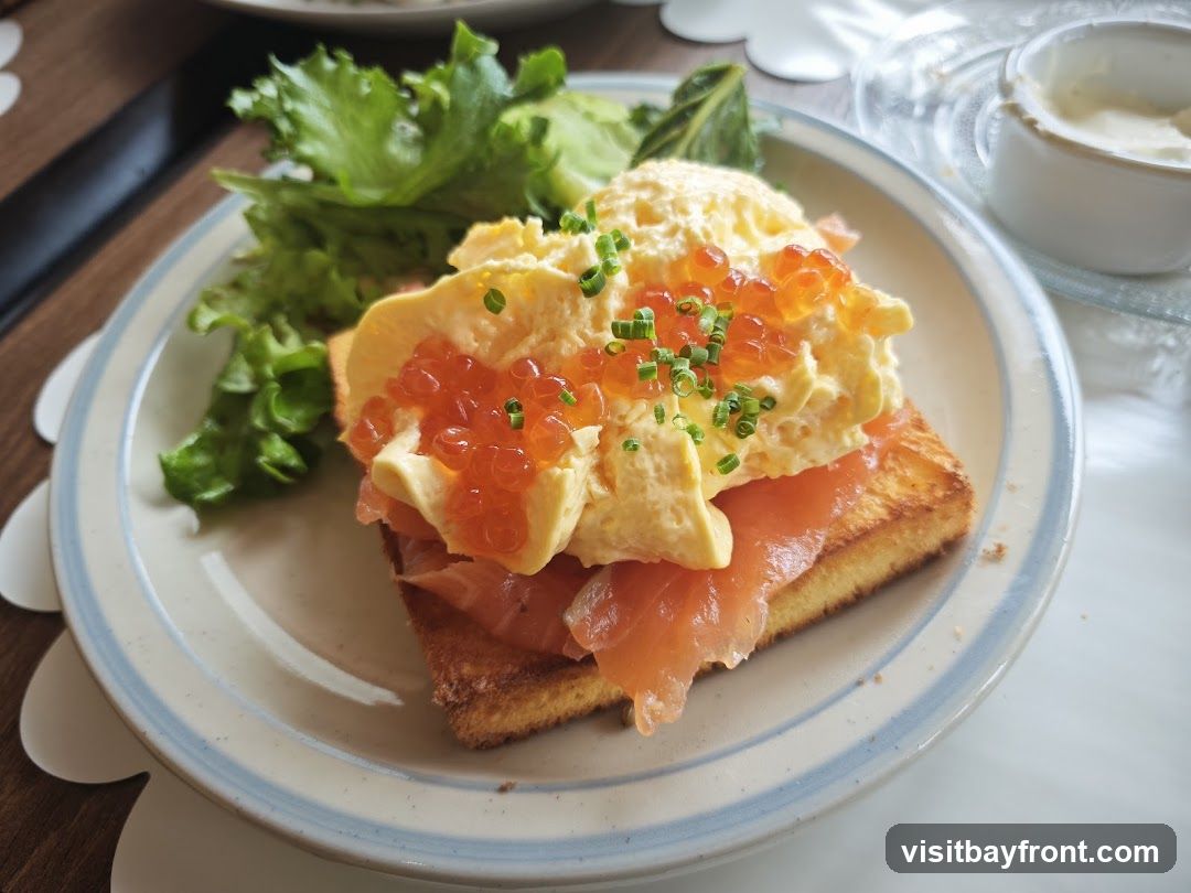 A close-up shot of a piece of toast topped with smoked salmon, a generous portion of scrambled eggs, and bright orange salmon roe. The dish is garnished with chopped chives and served with a side of lettuce and a small dish of what appears to be a creamy sauce. The meal is presented on a plate with a blue and white striped rim.