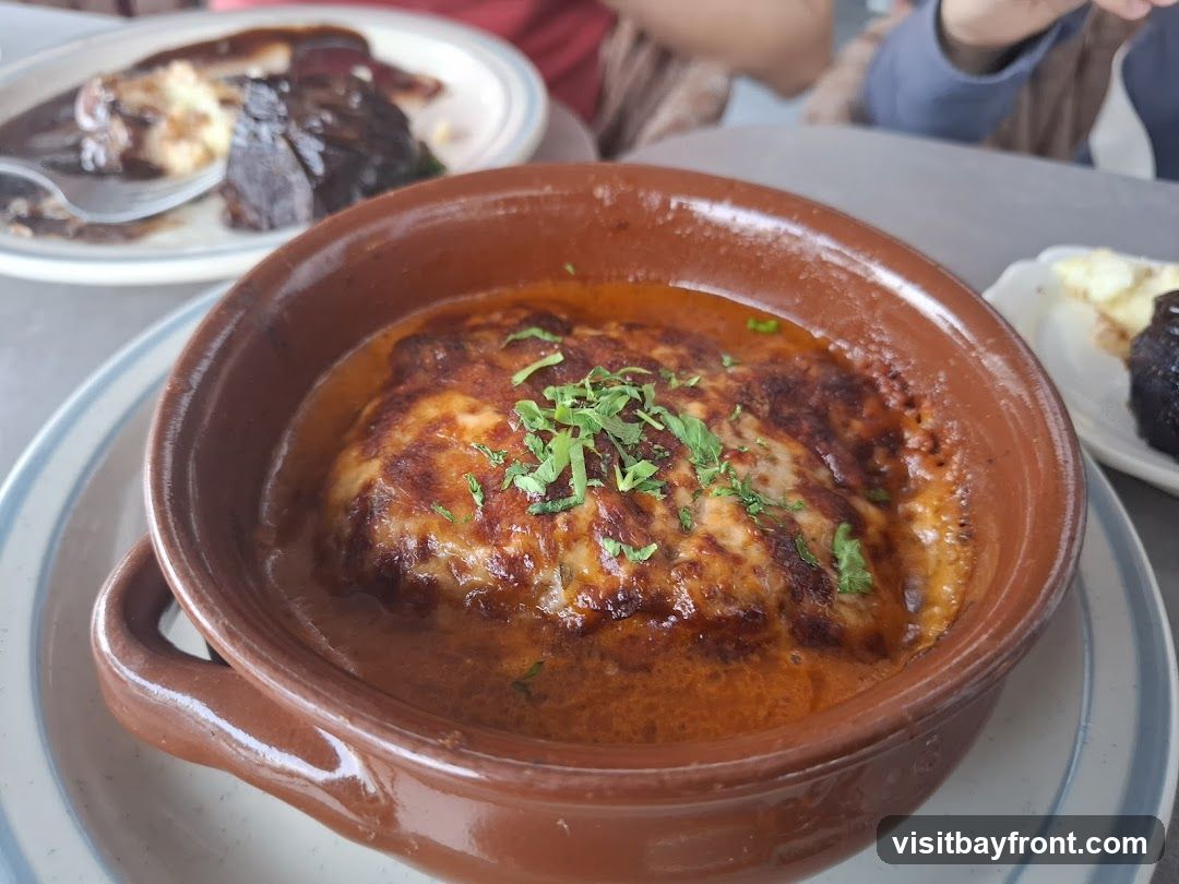 A close-up shot of a baked dish in a brown ceramic bowl. The dish is covered in melted, browned cheese and sprinkled with chopped green herbs. It is served in a rich, orange-red sauce. In the background, out of focus, are other plates of food, including one with what appears to be meat in a dark sauce and mashed potatoes, and another with a dark, possibly fried item and some white garnish. The setting appears to be a restaurant.