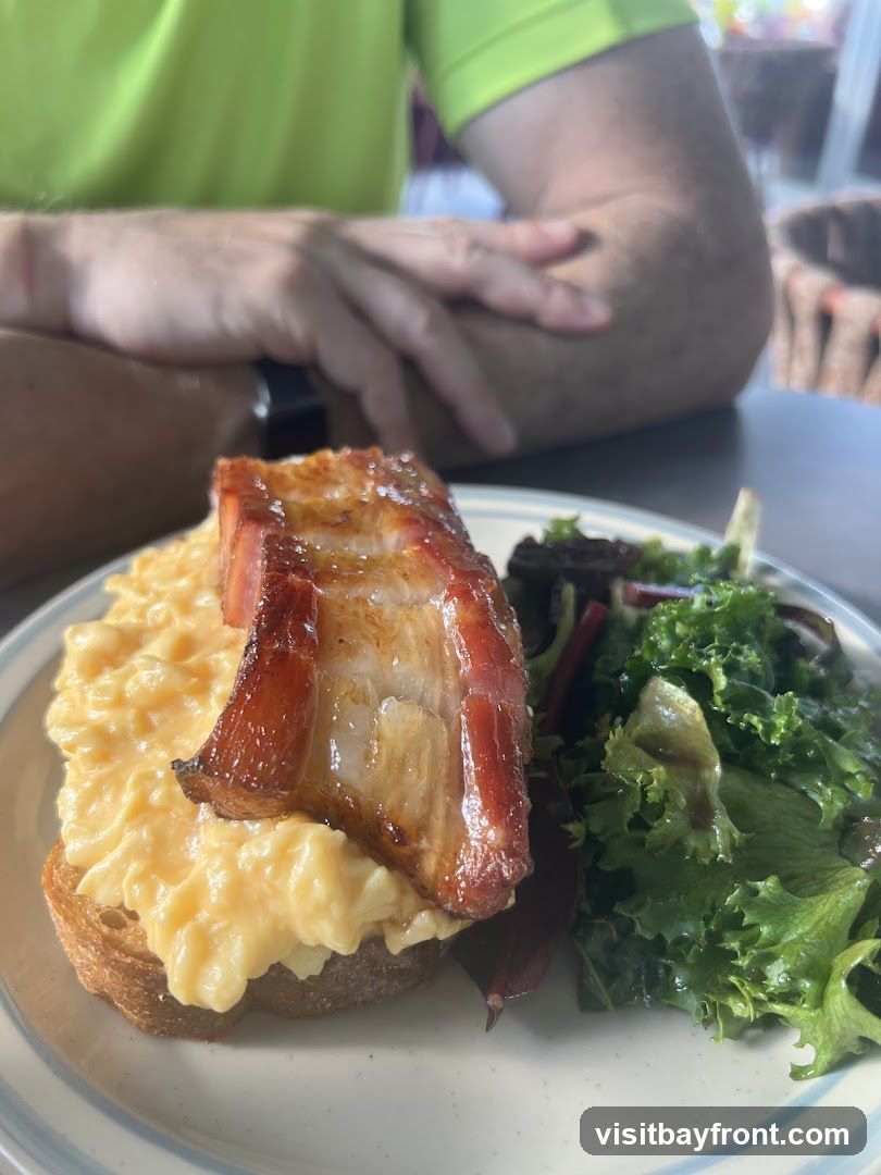 A close-up shot of a dish featuring scrambled eggs piled high on a slice of toast, topped with a thick piece of bacon. A small side salad with mixed greens is served alongside. A person wearing a lime green shirt is visible in the background, with their arms crossed.