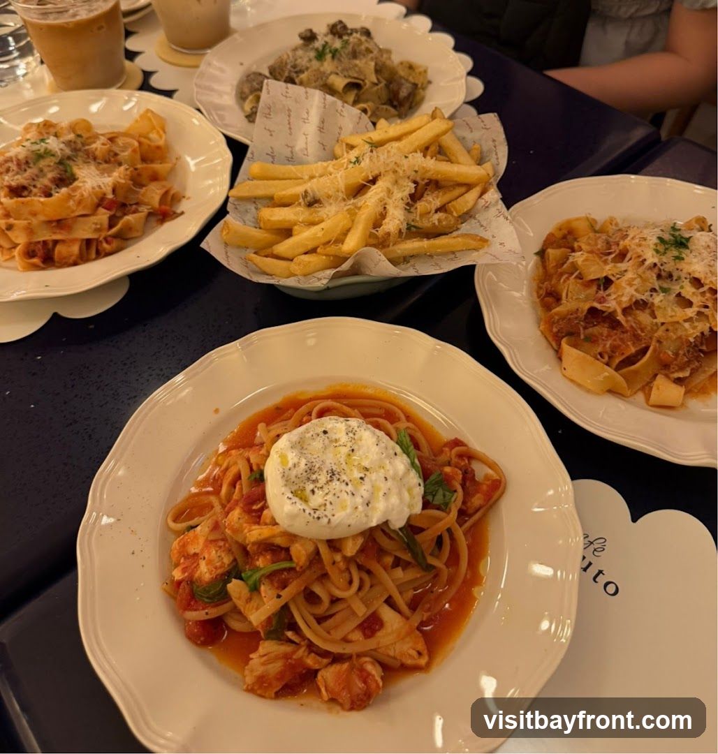 A table laden with several plates of pasta and a basket of french fries. In the foreground, a plate of linguine with a rich tomato sauce, chunks of chicken, and a dollop of burrata cheese topped with black pepper and olive oil. To the right, a plate of pappardelle pasta with a meat sauce. In the background, another plate of pasta with a creamy sauce and mushrooms, and a basket of golden french fries sprinkled with grated cheese. Two drinks, possibly iced coffee or tea, are visible in the upper left corner. A white paper placemat with a logo that reads "Cafe ...uto" is partially visible on the right.