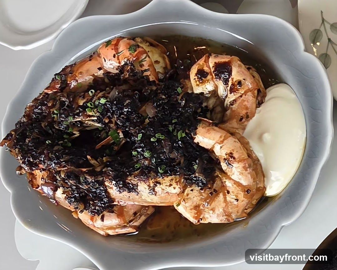 A close-up overhead view of a dish featuring several large, grilled shrimp served in a light brown sauce. The shrimp are topped with a dark, finely chopped mixture, possibly crispy garlic or shallots, and garnished with chopped green onions. A dollop of white sauce, likely aioli or mayonnaise, is on the side. The dish is presented in a light gray, scalloped-edge bowl.