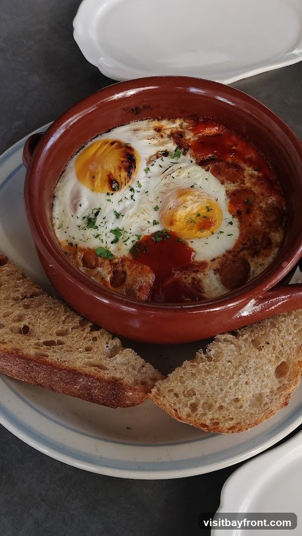 A close-up shot of baked eggs in a rustic brown dish, topped with a red sauce and sprinkled with parsley. Two slices of crusty bread are placed on the side of the dish, on a light-colored plate with a blue rim. The dish appears to be served on a dark gray surface.