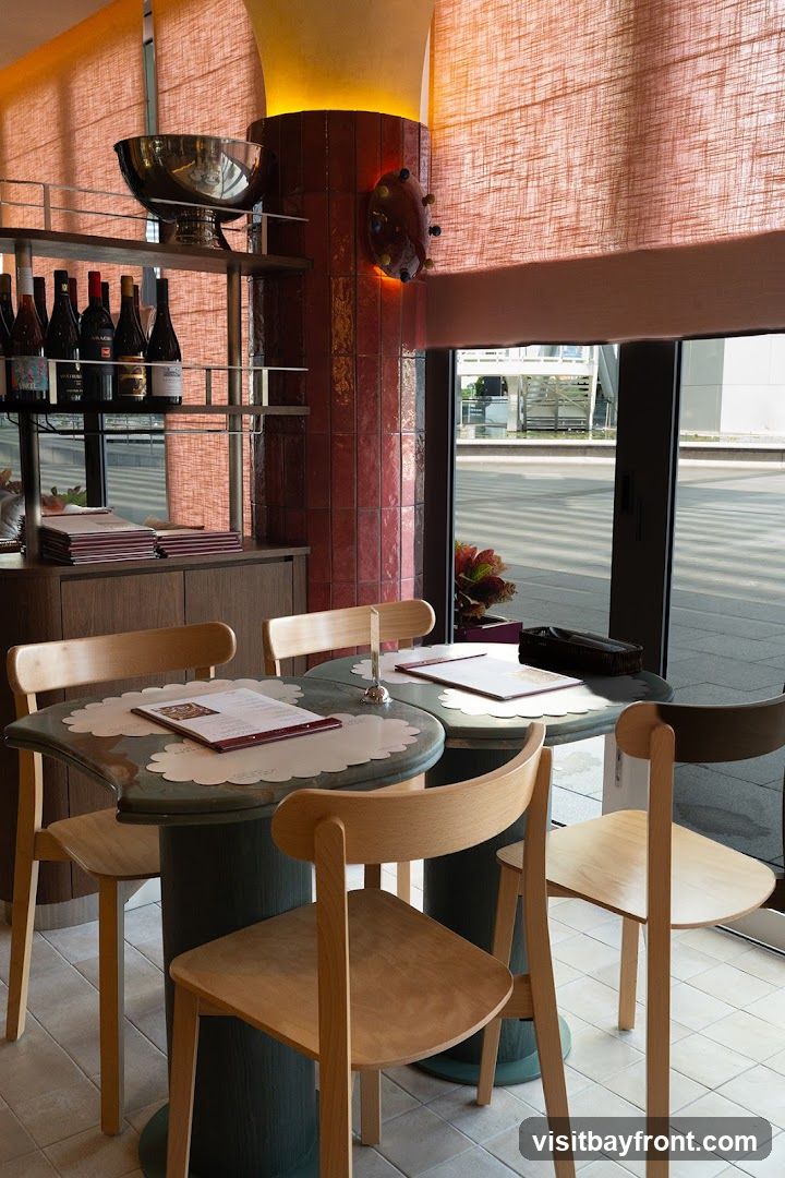 The image shows the interior of a restaurant with several tables set for dining. Wooden chairs surround the tables, which are covered with placemats and menus. In the background, shelves hold several bottles of wine, and a large silver bowl is displayed. A red tiled pillar with a decorative light fixture is visible. The overall ambiance is modern and clean, with natural light coming in from a large window.
