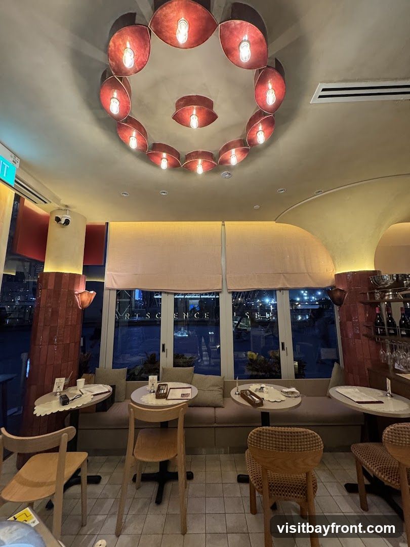 The interior of a restaurant with tables and chairs set up for dining. A large circular light fixture hangs from the ceiling. Large windows offer a view of the outside at night.