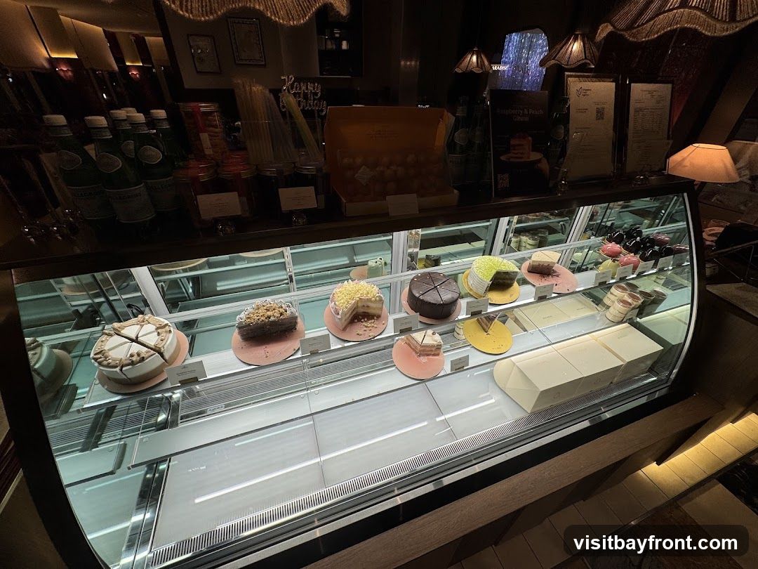 A refrigerated display case filled with various cakes and pastries. Bottles of San Pellegrino water are visible on the shelf above. There are also jars of ingredients and a box of what appears to be macarons. A sign with "Happy Birthday" is also present.