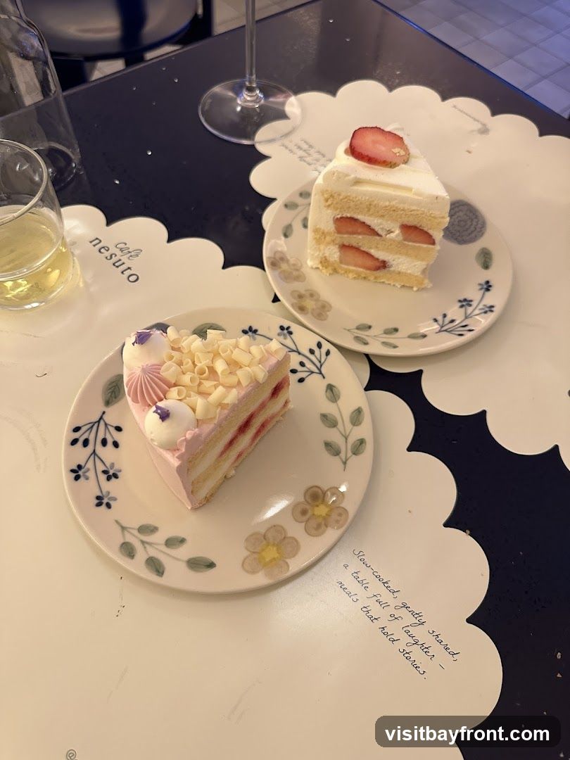 Two slices of cake are on decorative plates. One slice is a strawberry shortcake, and the other is a pink layered cake topped with white chocolate shavings and a purple flower decoration. A glass of light-colored liquid and a wine glass are also visible on the table. A quote about shared meals is written on a placemat.