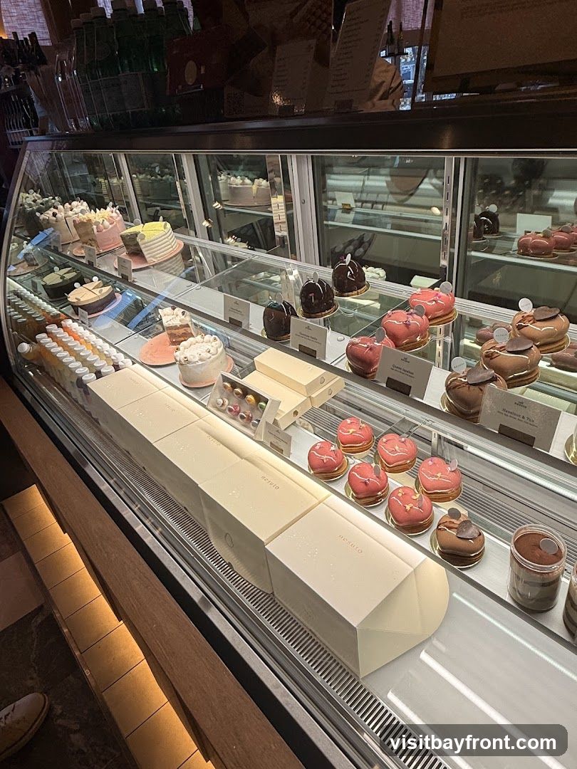 A refrigerated display case filled with various cakes, pastries, and desserts. The desserts include whole cakes, individual tarts, and mousse-like creations, many decorated with fruit and chocolate. Boxes with the brand name 'nesuto' are also visible.