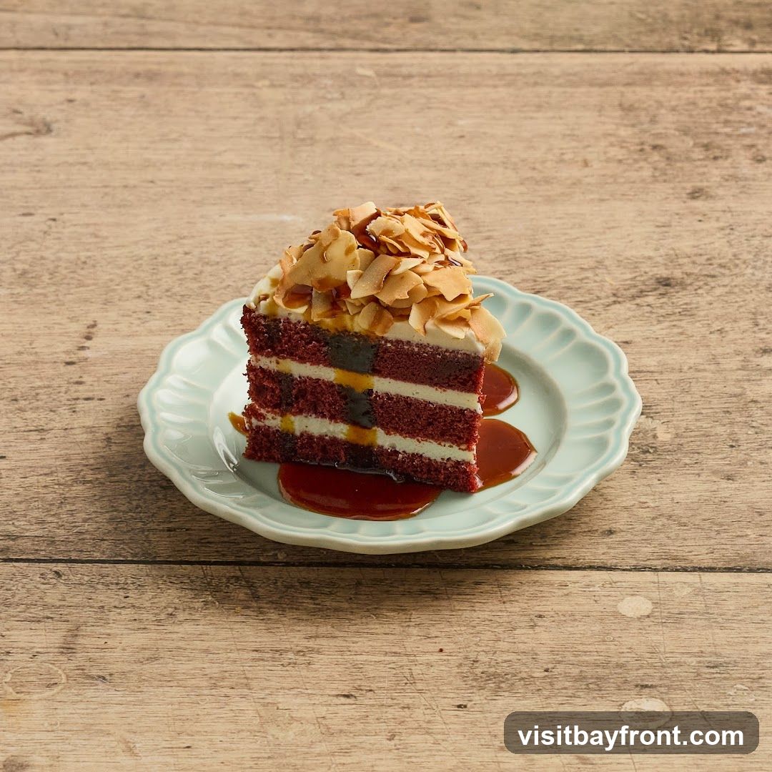 A slice of red velvet cake with white frosting and a dark filling, topped with toasted coconut flakes and drizzled with caramel sauce, served on a light blue scalloped plate against a wooden background.