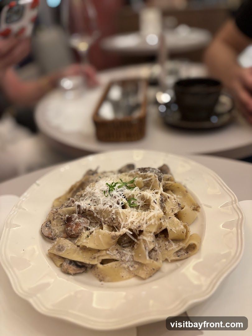 A close-up shot of a plate of pappardelle pasta with a creamy mushroom sauce, topped with grated cheese and a sprig of garnish. The dish is served on a white, scalloped-edge plate. In the blurred background, people are seated at tables, with a cup of coffee and a small woven basket visible.