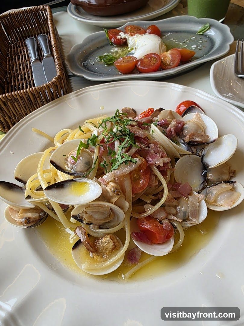 A close-up shot of a plate of spaghetti with clams, cherry tomatoes, and bacon. The pasta is coated in a light, oily sauce. In the background, a dish of burrata cheese with tomatoes and basil is visible, along with a basket of cutlery.