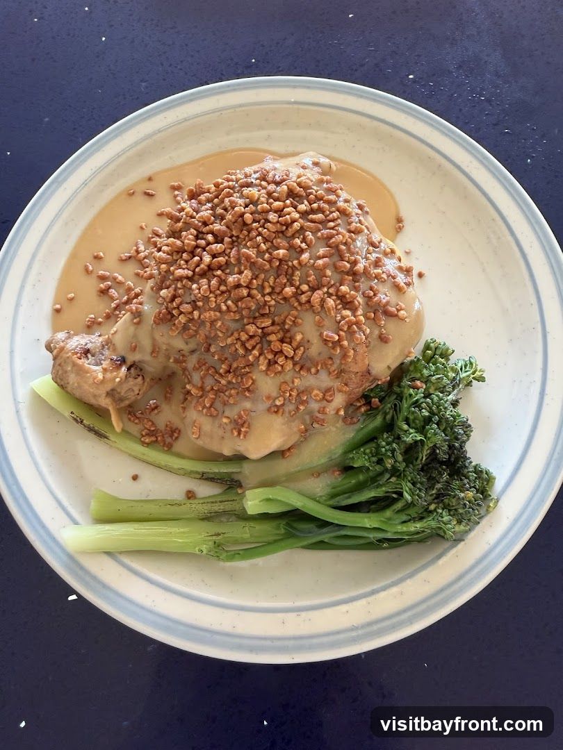 A close-up overhead shot of a plate of food. The dish features a piece of chicken covered in a creamy sauce and sprinkled with small, crunchy toppings. Alongside the chicken are several stalks of broccolini. The plate has a blue and white striped rim and is set against a dark blue surface.