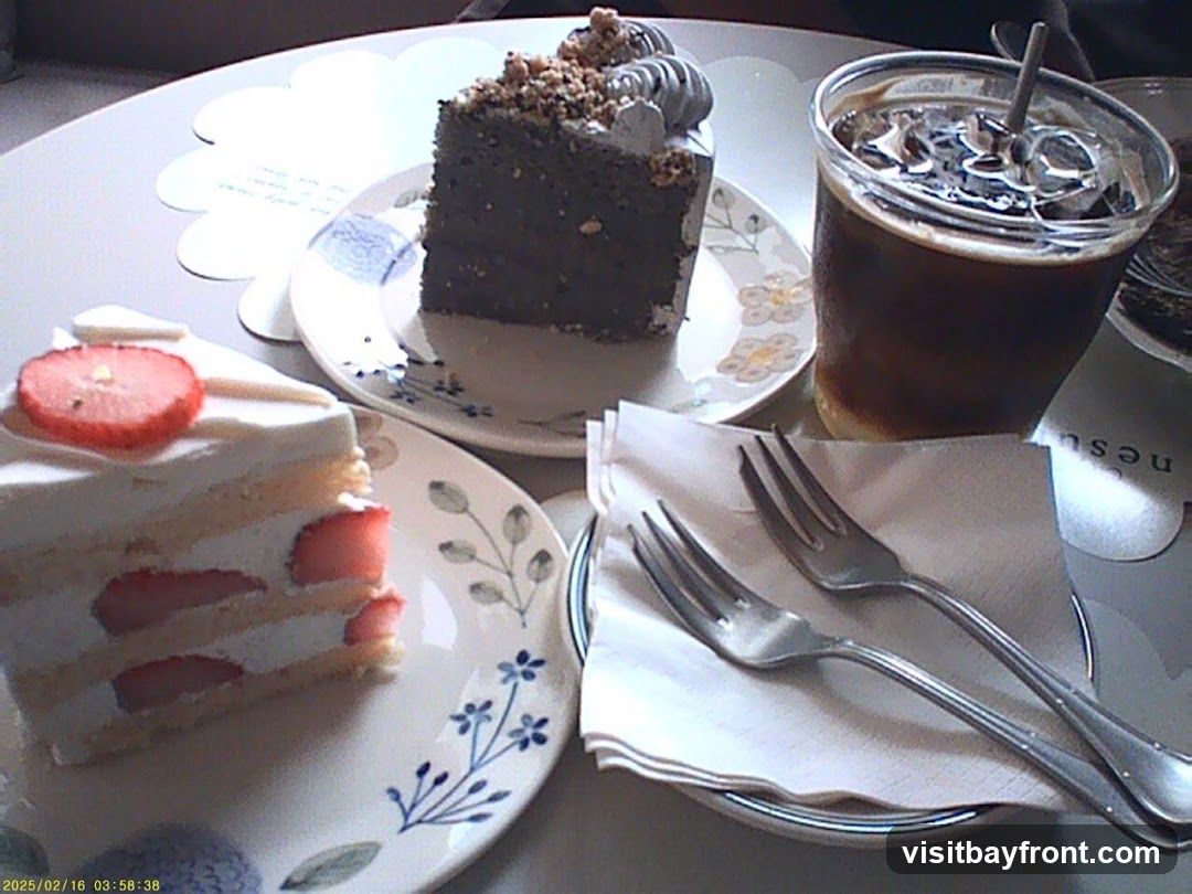 A slice of strawberry shortcake and a slice of dark cake are on plates on a table. A glass of iced coffee is also on the table, along with two forks on a napkin.