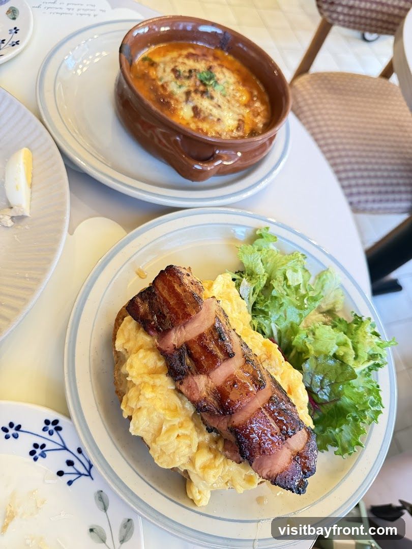 A plate of scrambled eggs topped with thick-cut bacon and a side of mixed greens. Another plate in the background holds a dish of baked pasta with cheese and sauce. The setting appears to be a cafe or restaurant with a light-colored table and chairs visible.