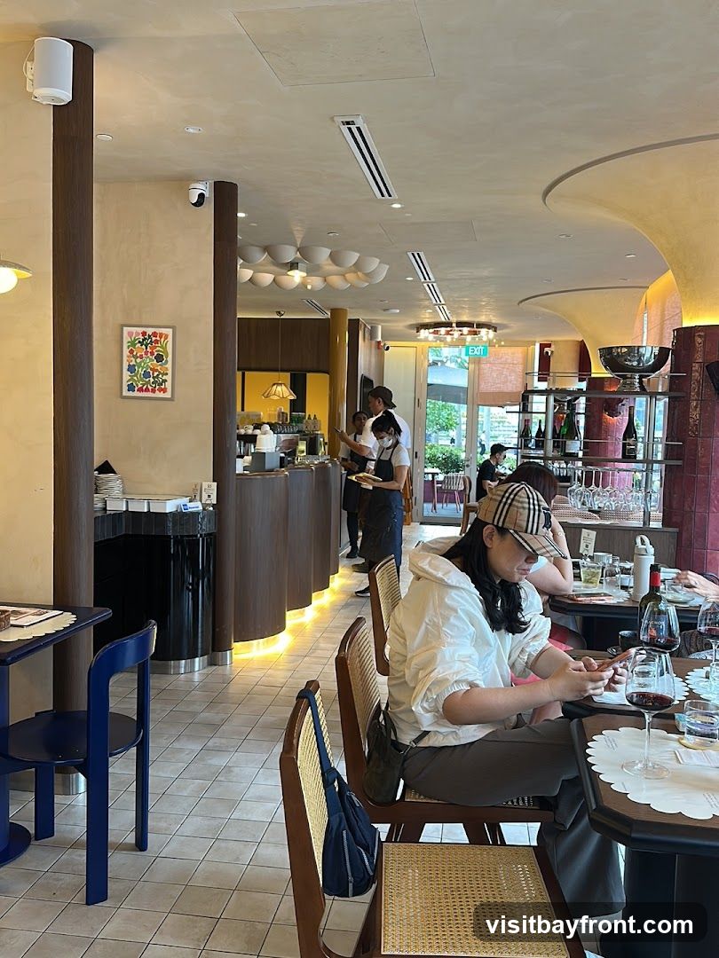 The image shows the interior of a restaurant with several patrons seated at tables. A woman in a white jacket and a checkered cap is looking at her phone. Staff members are visible behind a counter. The decor includes modern lighting fixtures and artwork on the wall.