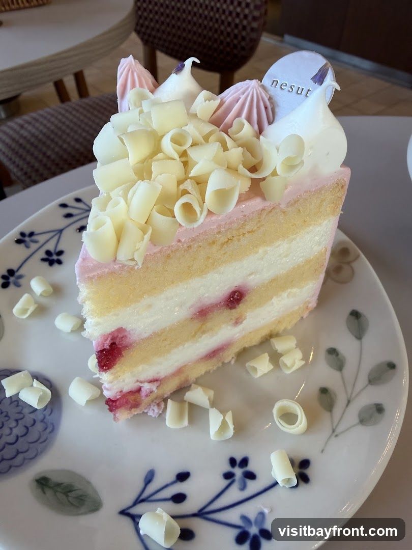 A slice of layered cake with pink frosting and white chocolate shavings is presented on a decorative plate. The cake has visible layers of sponge cake and cream filling, with hints of red fruit. The plate features a blue floral design. A small circular logo with the text "nesut" is visible on top of the cake.