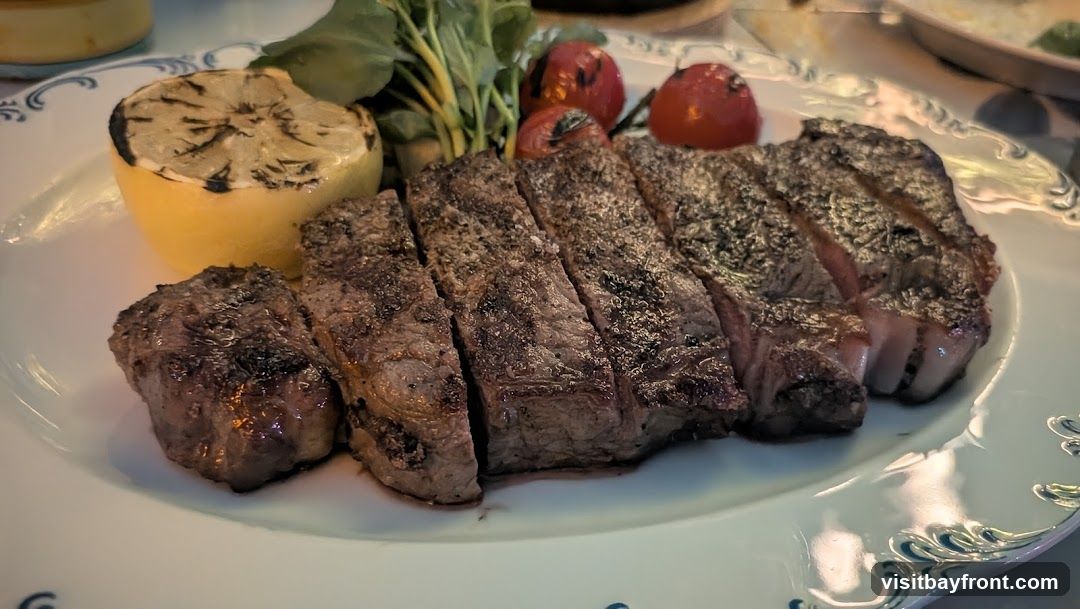 A plate of sliced steak, grilled lemon, cherry tomatoes, and greens. The steak is cooked to medium-rare and seasoned with salt and pepper. The plate has a decorative blue and white trim.