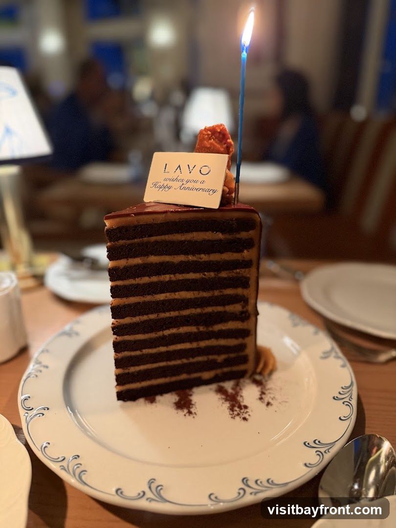 A tall slice of chocolate cake with multiple layers of chocolate cake and caramel frosting is served on a decorative white plate. A lit candle is placed on top of the cake, next to a small card that reads "LAVO wishes you a Happy Anniversary." The background is blurred, showing a restaurant interior with diners.