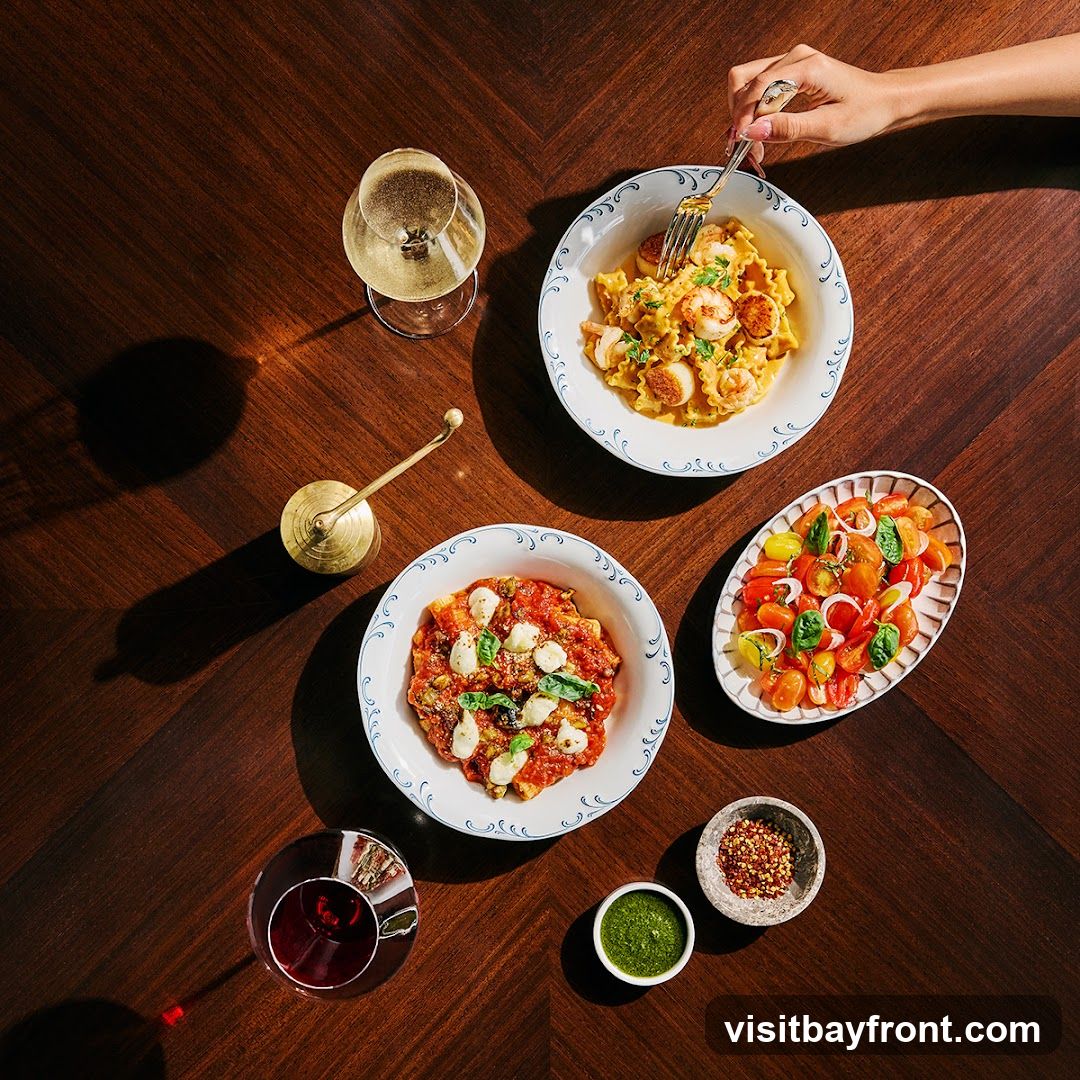 A top-down view of a wooden table set with a meal. In the center, a bowl of pasta with scallops and a creamy sauce is being eaten with a fork. To the left, a bowl of pasta with tomato sauce and mozzarella balls is visible, alongside a glass of red wine. To the right, a salad of cherry tomatoes and onions is presented in an oval dish. Small bowls of green sauce and red pepper flakes are also on the table, along with a glass of white wine and a brass condiment holder.