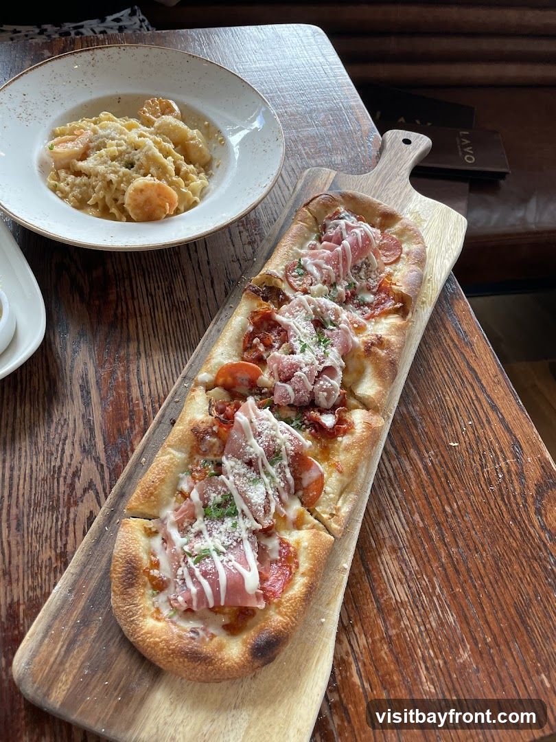 A flatbread topped with prosciutto, pepperoni, and cheese is served on a wooden board next to a bowl of pasta with shrimp and grated cheese. A menu with the word "LAVO" is visible in the background.