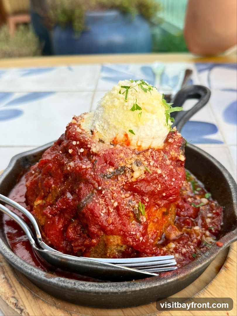 A large meatball covered in marinara sauce and topped with cheese and parsley, served in a cast-iron skillet with a fork and spoon. The dish is placed on a tiled surface with a blurred background of plants and pottery.