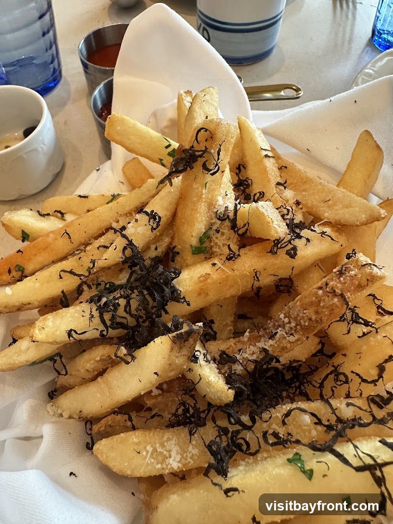 A close-up shot of a basket of truffle fries, seasoned with parmesan cheese and herbs. The fries are golden brown and crispy, served in a white cloth-lined basket. In the background, there are small metal cups with ketchup, a white ceramic dish, and blue glassware.