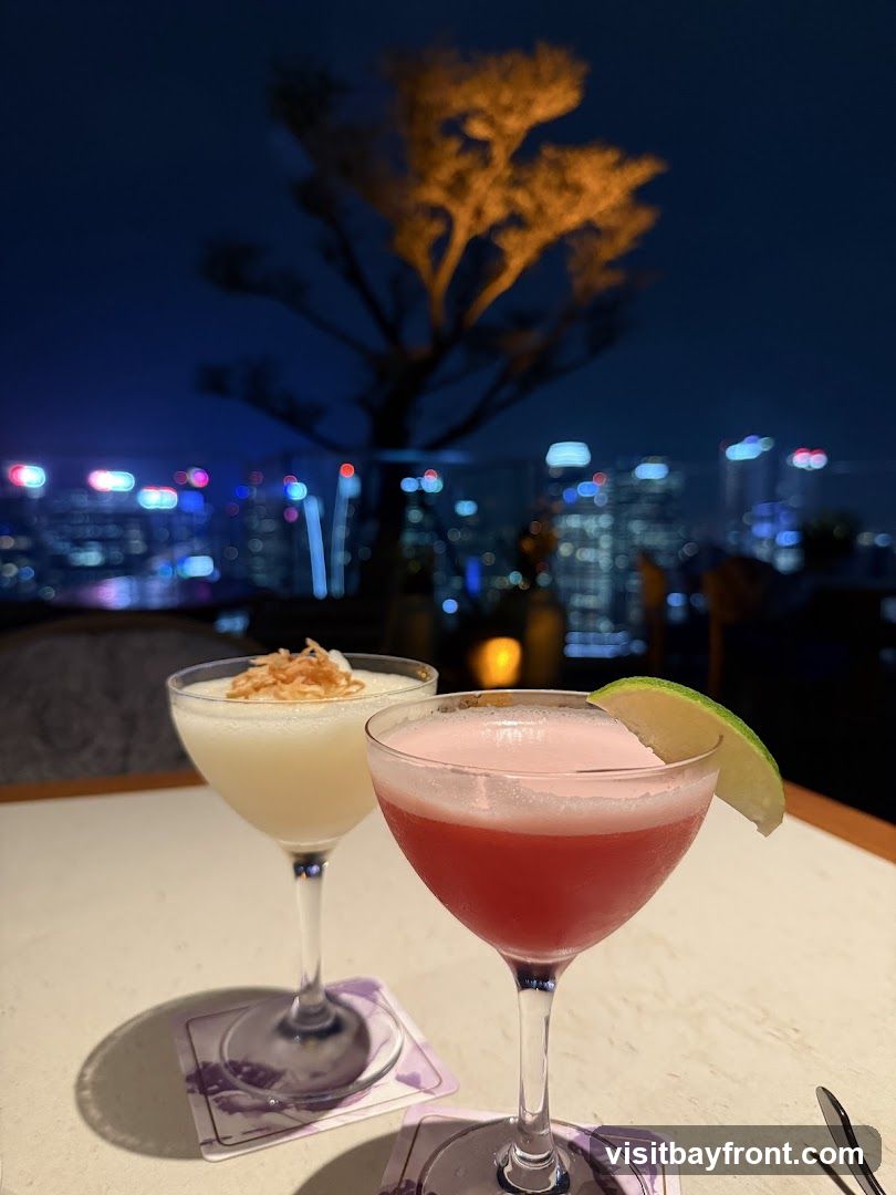 Two cocktails are on a table with a city skyline in the background at night. The cocktail on the left is white with a garnish of shredded coconut. The cocktail on the right is pink with a lime wedge garnish.