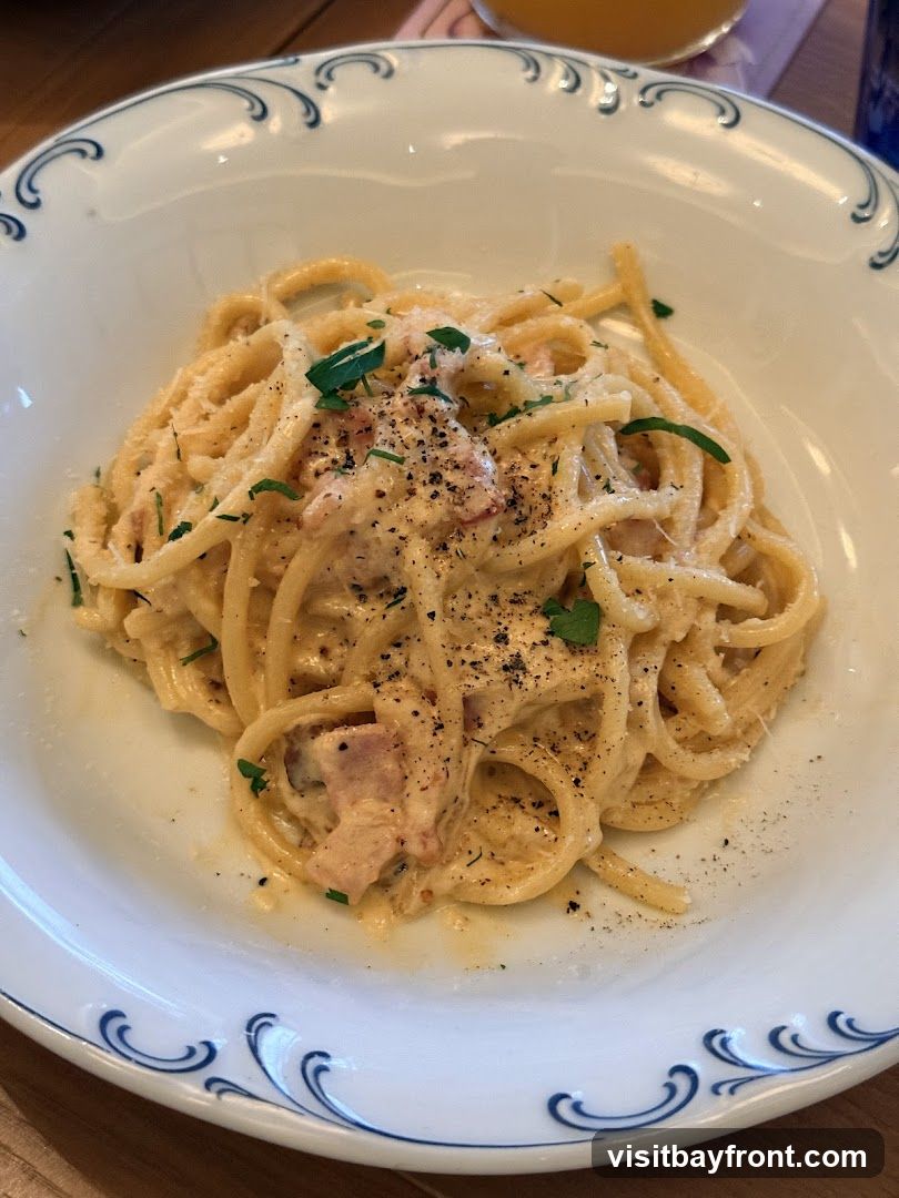 A close-up shot of a plate of spaghetti carbonara, garnished with parsley and black pepper. The pasta is coated in a creamy sauce with visible pieces of bacon. The plate has a decorative blue wave pattern around the rim.