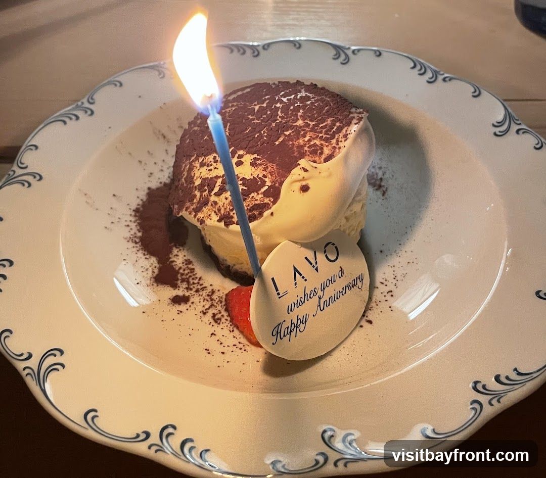 A dessert, possibly tiramisu, topped with cocoa powder and cream, is served on a decorative plate. A lit candle is stuck into the dessert, and a small oval plaque reads "LAVO wishes you a Happy Anniversary". A strawberry slice is placed next to the plaque.