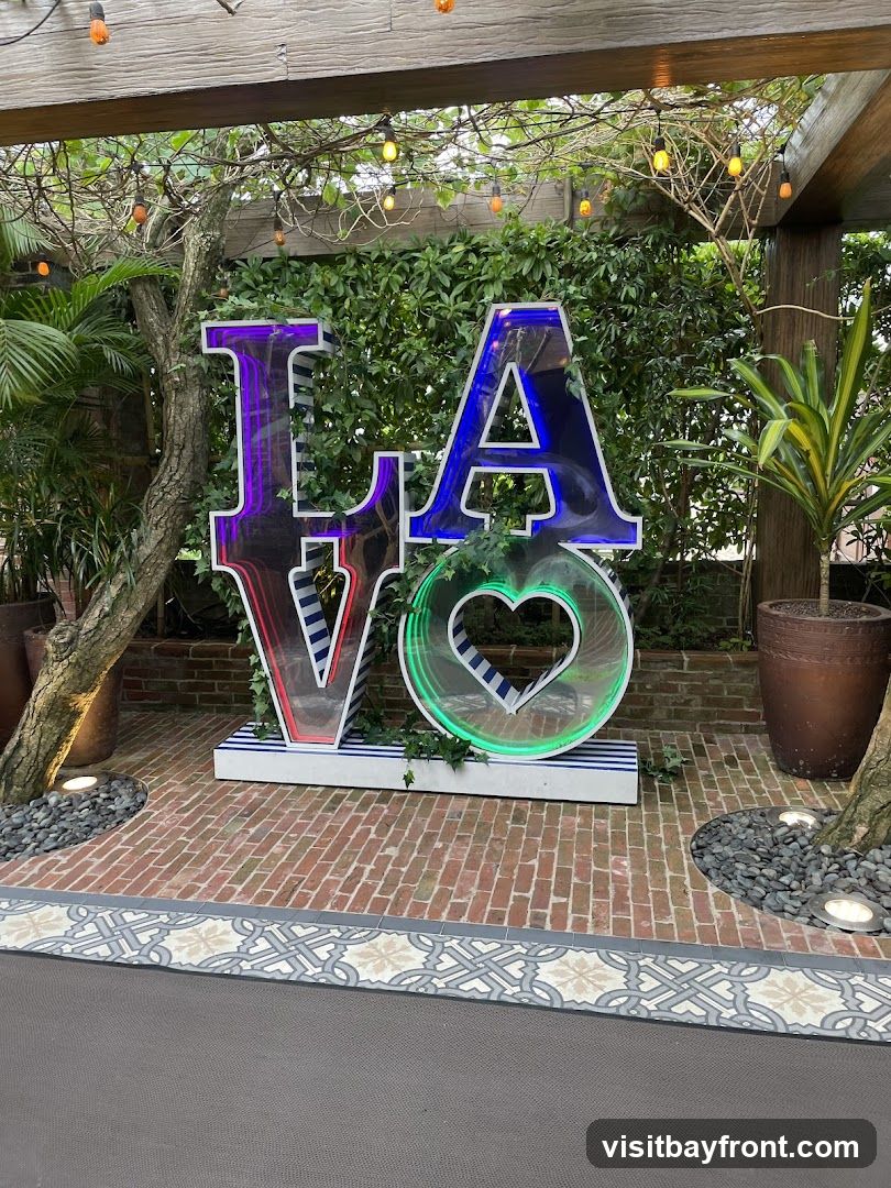 A large, illuminated sign spelling out "LA Love" is displayed outdoors. The letters are colorful, with purple and red for "L" and "A," and green for "Love" which is depicted as a heart shape. The sign is set against a backdrop of lush greenery and brickwork, with string lights overhead. Potted plants and decorative stones surround the base of the sign.