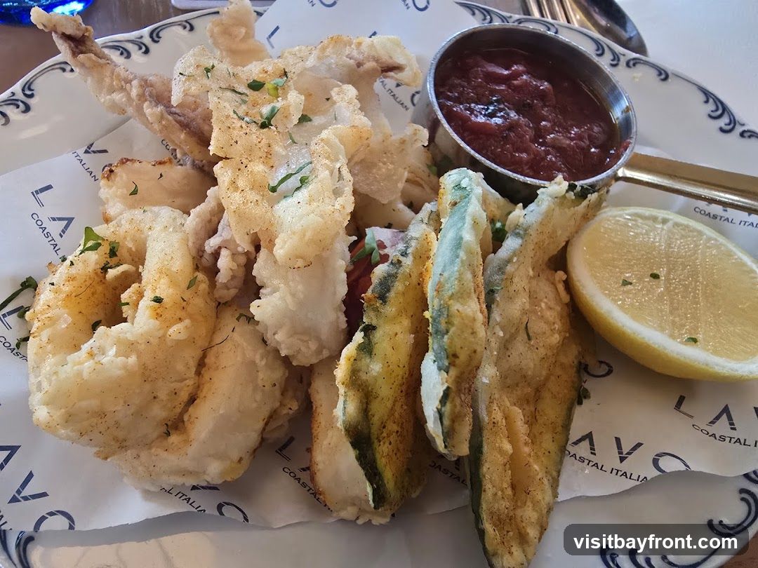 A plate of fried calamari and zucchini served with a side of marinara sauce and a lemon wedge. The food is presented on a white plate with a decorative blue rim, lined with paper featuring the logo 'LAVO COASTAL ITALIAN'.