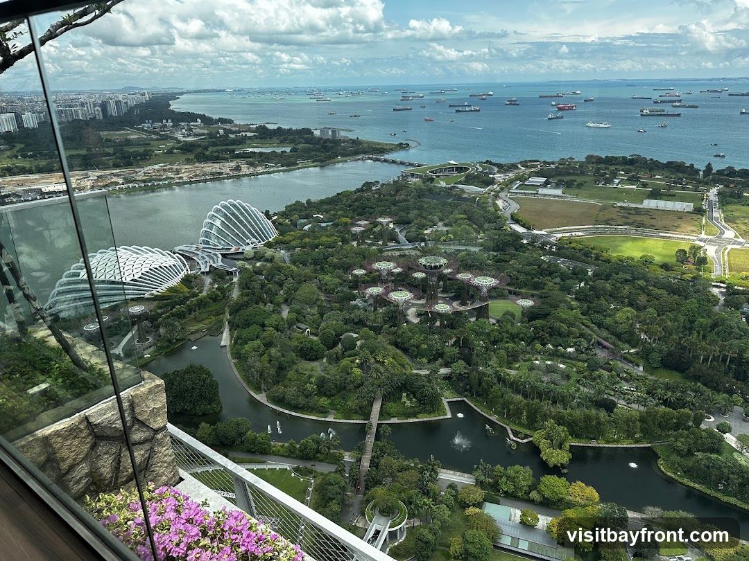 An aerial view of Gardens by the Bay in Singapore, showcasing the iconic Supertree Grove and the Cloud Forest and Flower Dome conservatories. The cityscape and a busy shipping lane in the bay are visible in the background. The perspective is from a high vantage point, possibly a building overlooking the gardens, with a balcony railing and purple flowers in the foreground.