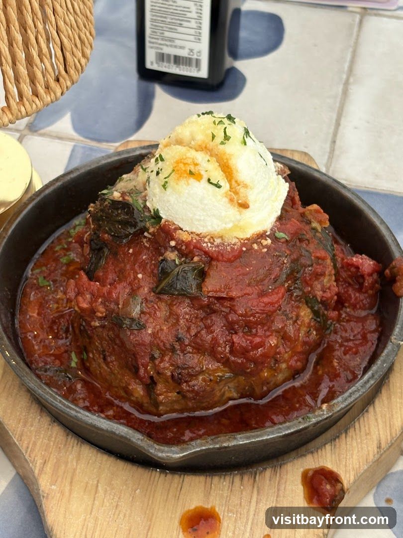 A large meatball is served in a cast-iron skillet, covered in a rich tomato sauce with visible herbs. A dollop of white cheese, possibly ricotta or mozzarella, is placed on top, sprinkled with parsley and a dusting of paprika. The skillet rests on a wooden board, and the background shows a tiled surface and a woven placemat.