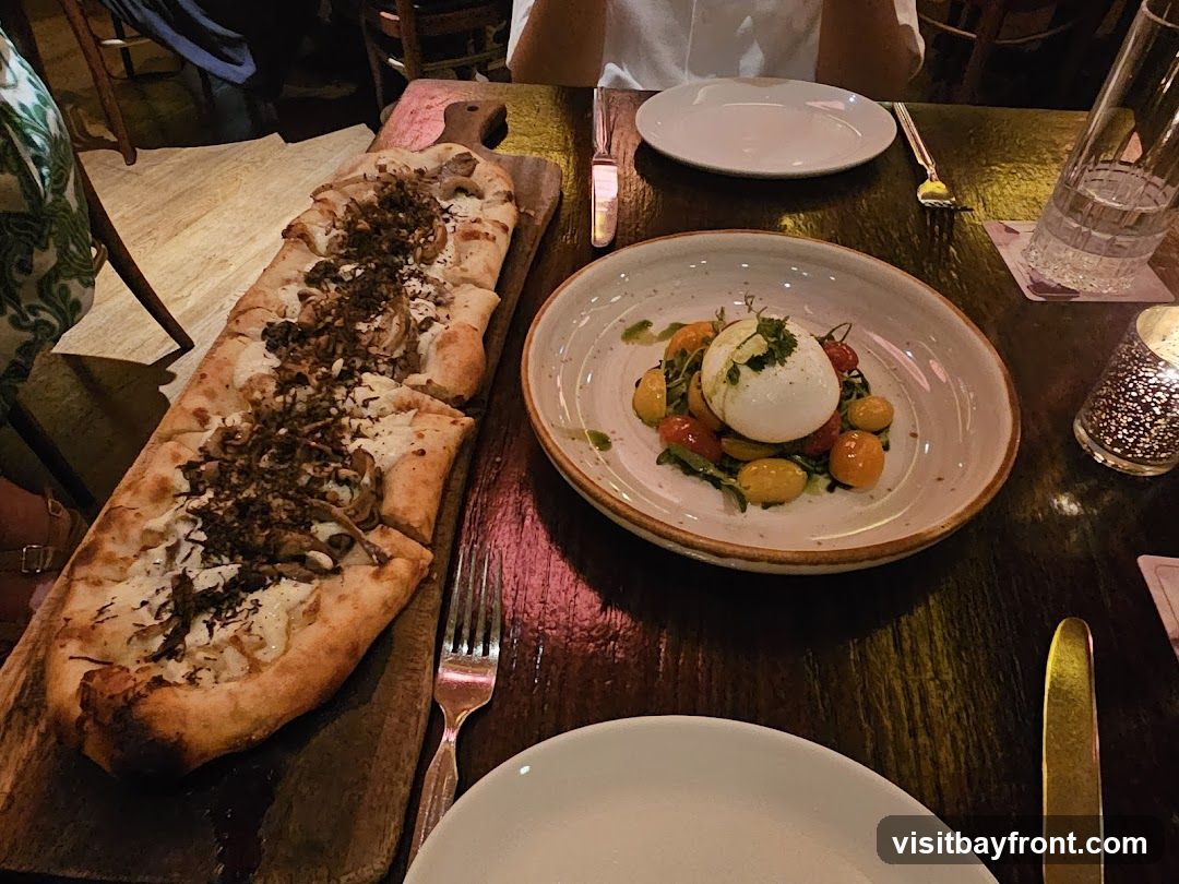 A long, thin flatbread topped with cheese and mushrooms is served on a wooden board. Next to it is a bowl of burrata cheese with cherry tomatoes and greens. The dishes are on a dark wooden table, with plates, cutlery, and a glass of water visible.
