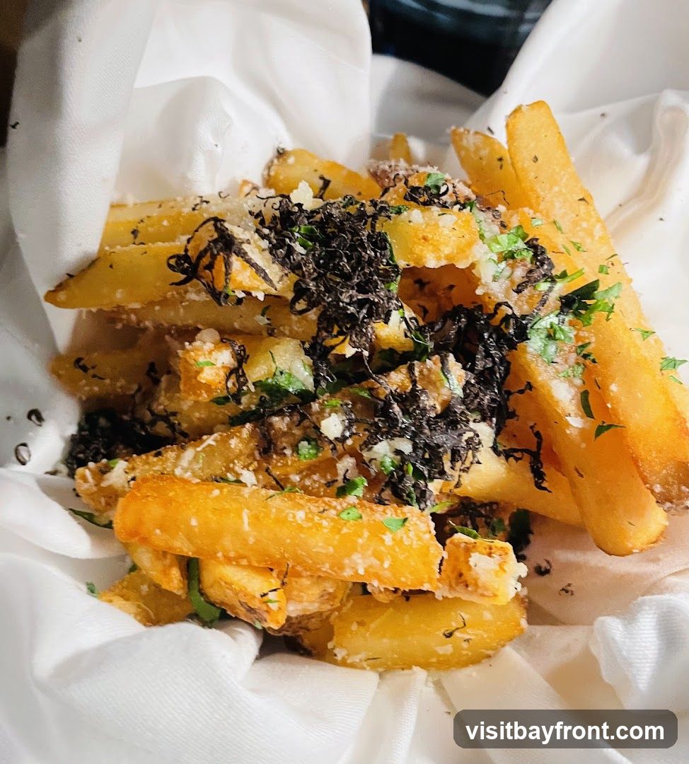 A close-up shot of truffle fries, generously topped with grated cheese, chopped parsley, and what appears to be truffle shavings. The fries are golden brown and served in a white cloth-lined basket.