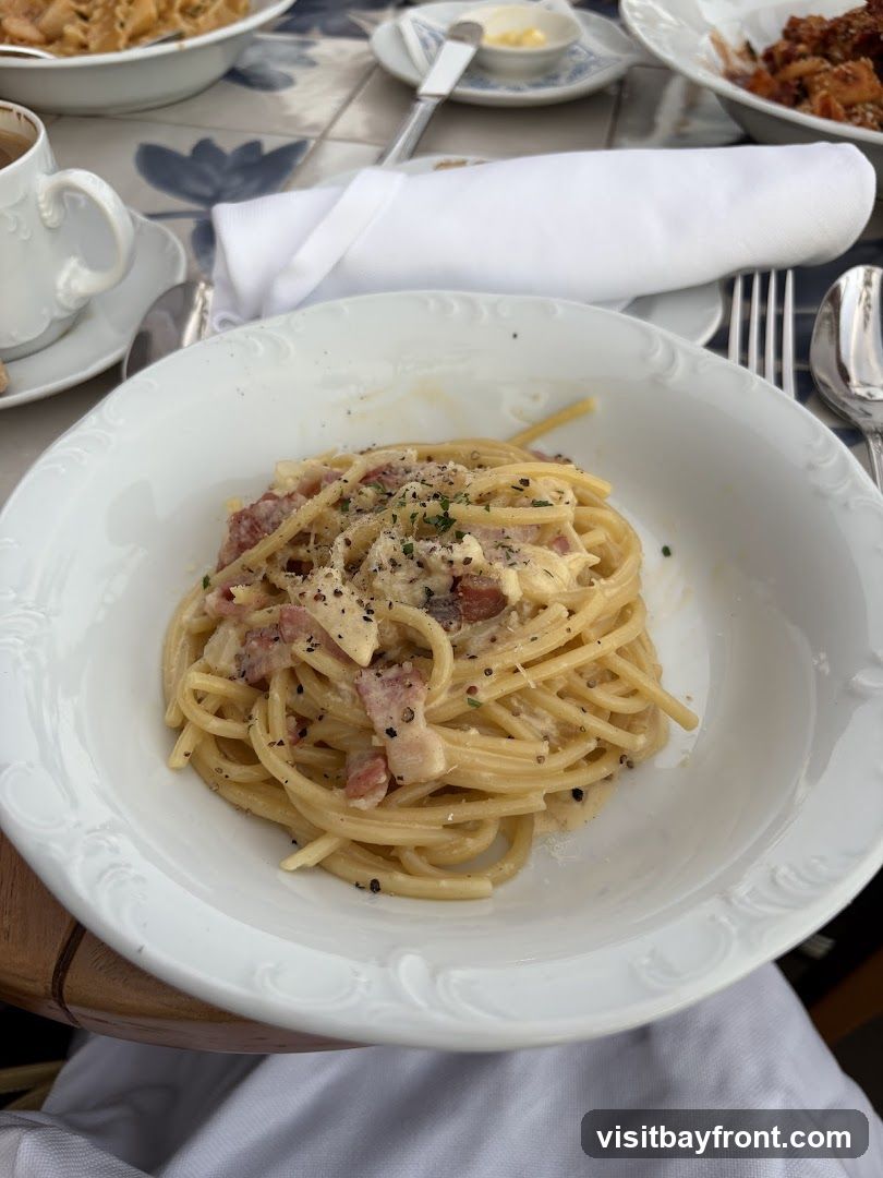A close-up shot of a bowl of spaghetti carbonara, garnished with parsley and black pepper. The pasta is coated in a creamy sauce with visible pieces of bacon or pancetta. In the background, other dishes and a cup of coffee are visible on a table with a patterned surface.