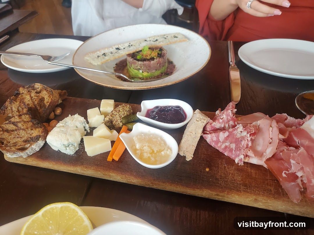 A wooden board filled with various meats, cheeses, bread, pickles, and two small dishes of preserves. A plate with a tuna tartare dish is visible in the background.