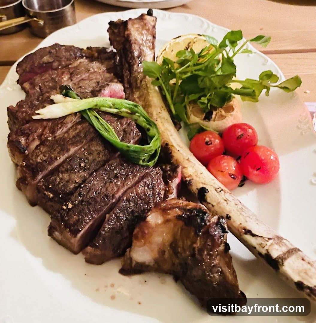 A large, sliced tomahawk steak is presented on a white plate, garnished with grilled lemon, roasted garlic, cherry tomatoes, and green onions. The steak is cooked to a medium-rare, with visible grill marks and seasoning. A small piece of bone is also on the plate.