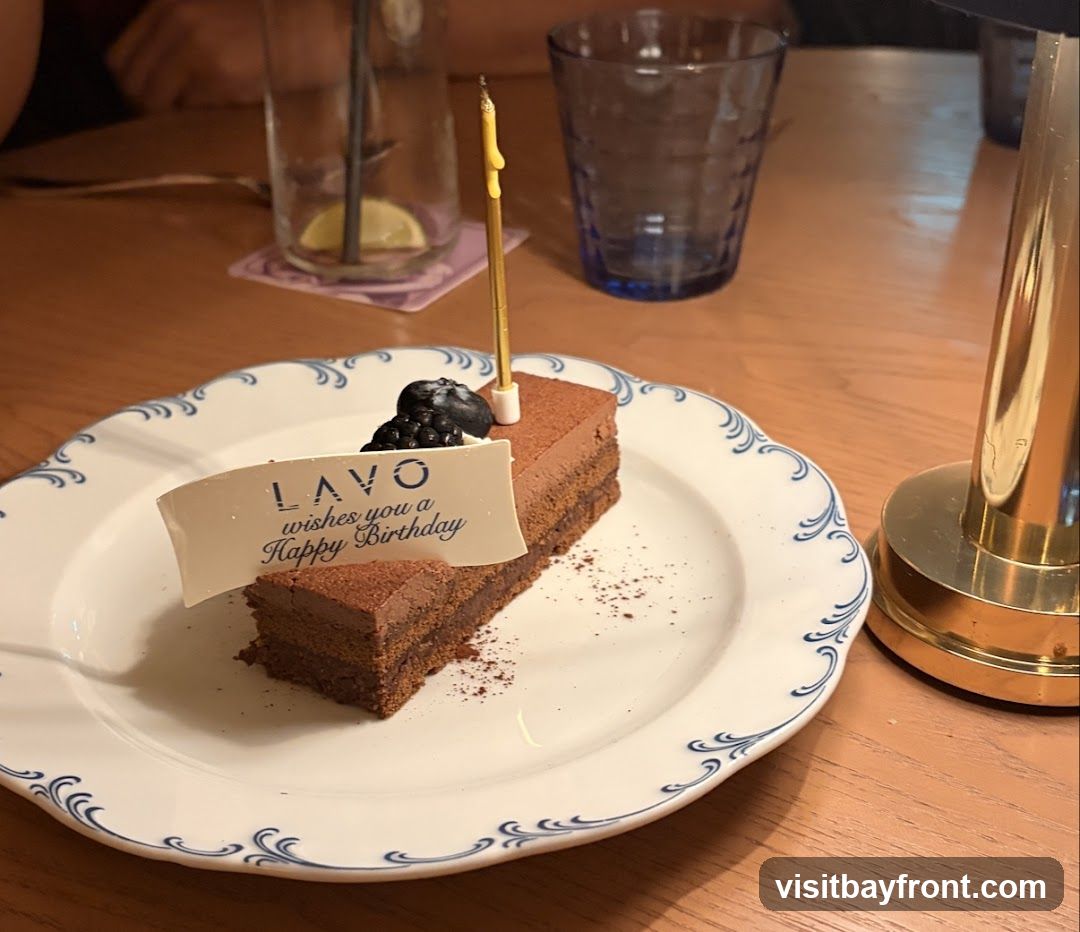 A slice of chocolate cake with a birthday candle and a blueberry on top is served on a white plate with blue trim. A white banner on the cake reads "LAVO wishes you a Happy Birthday." The cake is on a wooden table with glasses and a lamp in the background.