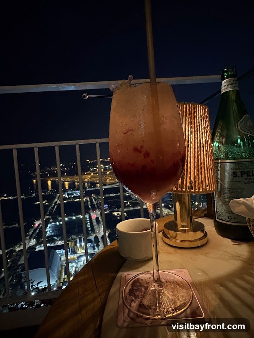 A tall glass of a red and orange layered cocktail with crushed ice and a metal straw sits on a table overlooking a city at night. A bottle of San Pellegrino sparkling water and a small lamp are also on the table.