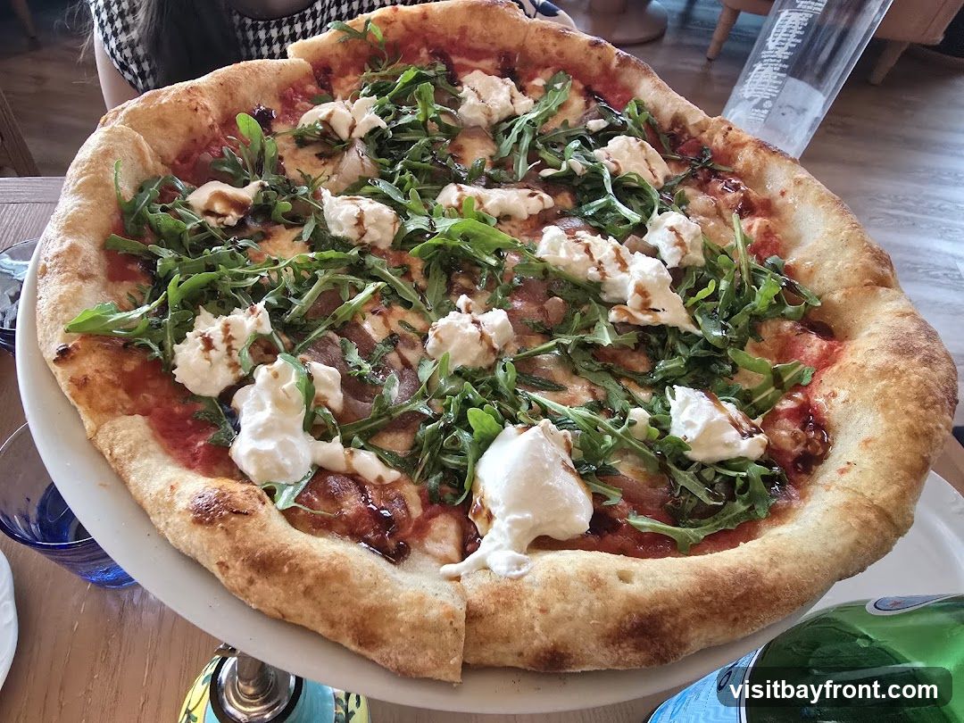 A close-up shot of a large pizza topped with fresh arugula, dollops of ricotta cheese, and a balsamic glaze. The pizza has a golden-brown crust and is presented on a white platter. In the background, a person wearing a black and white patterned top is visible, along with a green bottle of sparkling water. The setting appears to be a restaurant with wooden tables.