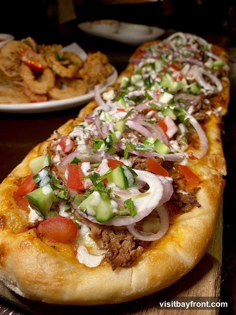 A long, flatbread pizza-like dish topped with shredded meat, diced tomatoes, cucumbers, red onions, and a white sauce is presented on a wooden board. In the background, a plate of fried calamari rings and other fried appetizers is visible.
