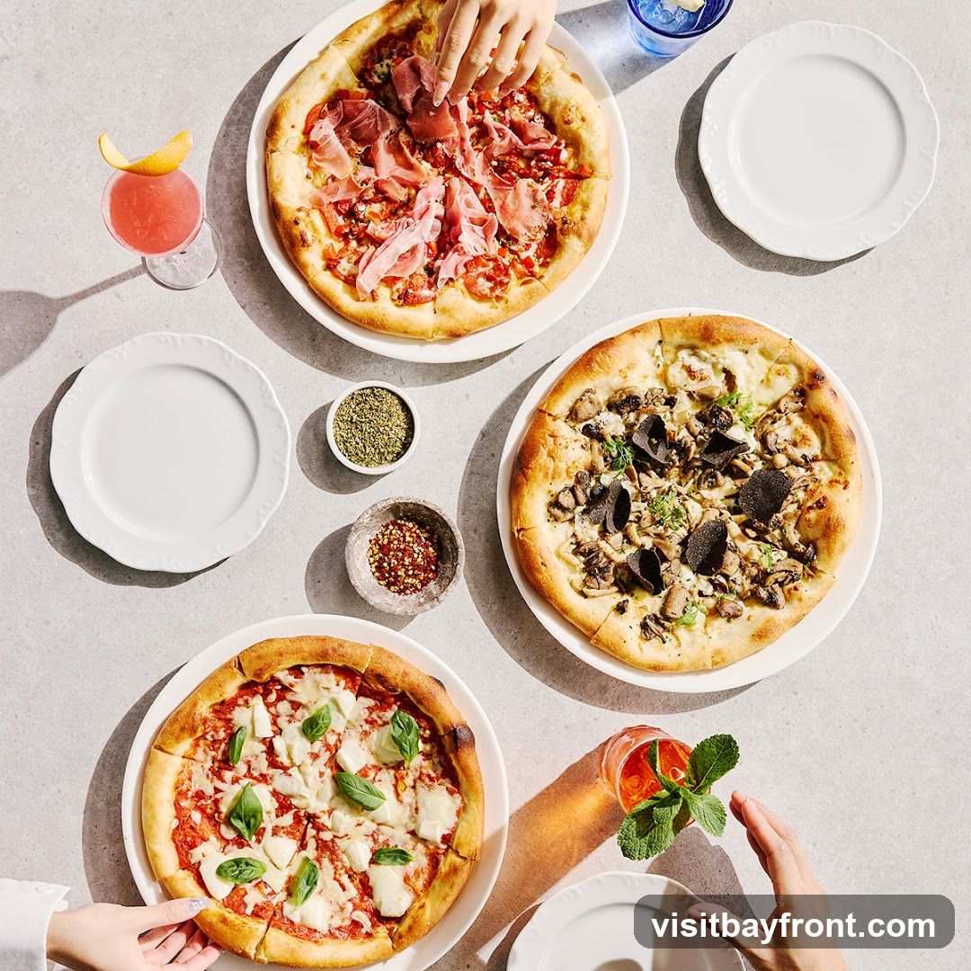 A top-down view of a table set with three pizzas, two cocktails, plates, and small bowls of spices. One pizza is topped with prosciutto and red sauce. Another pizza is topped with mushrooms, truffles, and cheese. The third pizza is a margherita with mozzarella, tomato sauce, and basil.