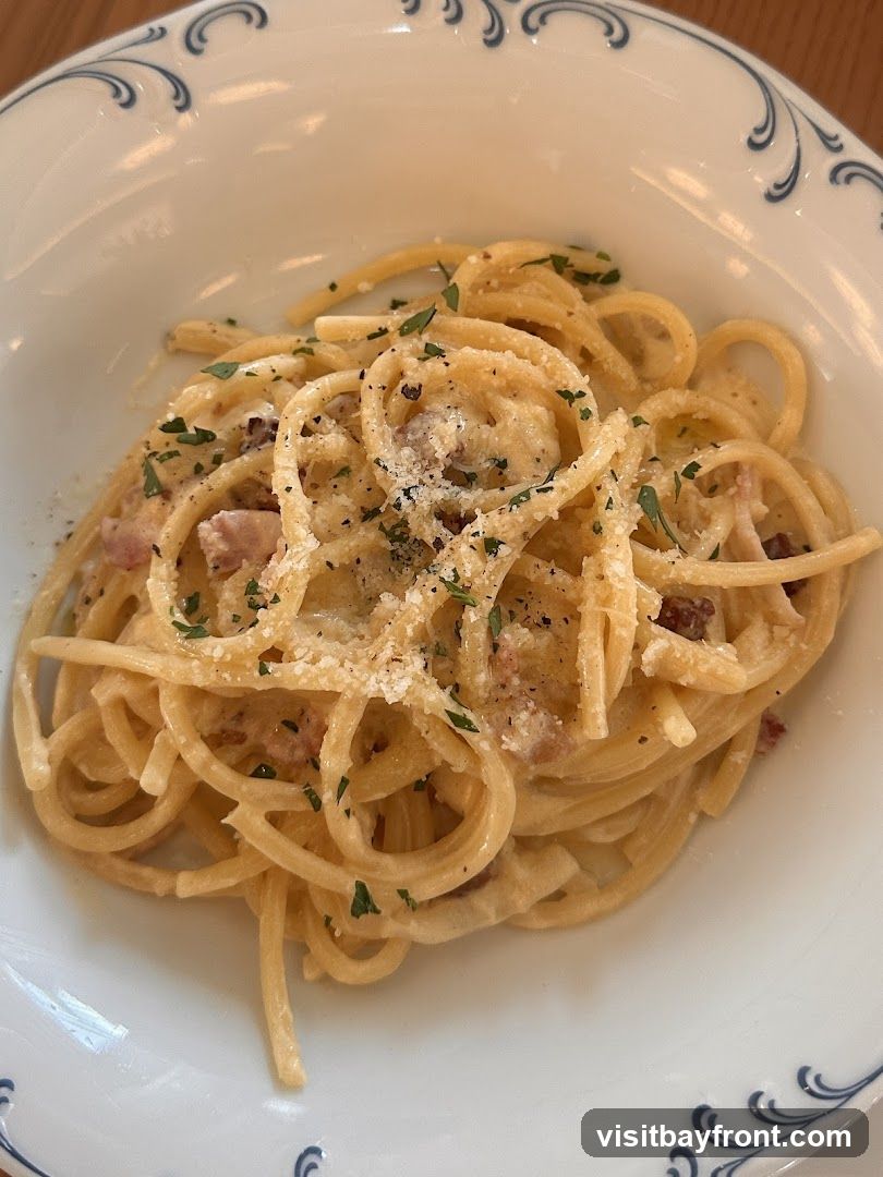 A close-up shot of spaghetti carbonara served in a white bowl with a decorative blue rim. The pasta is coated in a creamy sauce with visible pieces of pancetta and is sprinkled with grated cheese and chopped parsley.