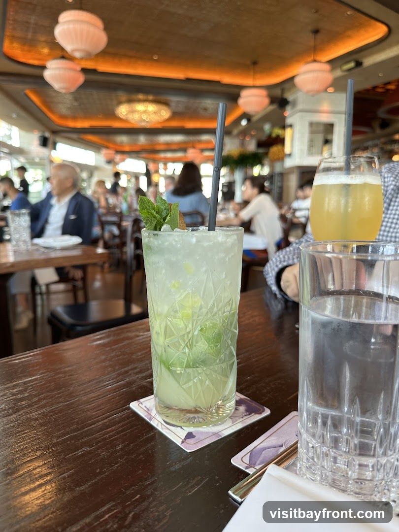 A close-up shot of two cocktails on a wooden table in a restaurant. The drink in the foreground is a mojito with mint and lime. In the background, other patrons are visible, along with decorative lighting fixtures on the ceiling.