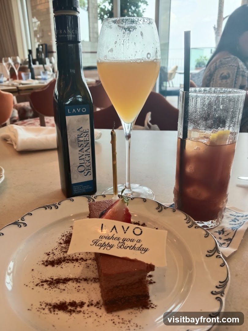 A slice of chocolate cake with a strawberry on top sits on a decorative plate. A white banner on the cake reads "LAVO wishes you a Happy Birthday." Next to the cake are two drinks: one in a champagne flute and another in a tall glass with a straw and lemon slice. A bottle of olive oil labeled "LAVO Olivastra Segginese" is also on the table. The background shows a restaurant interior with tables and chairs.