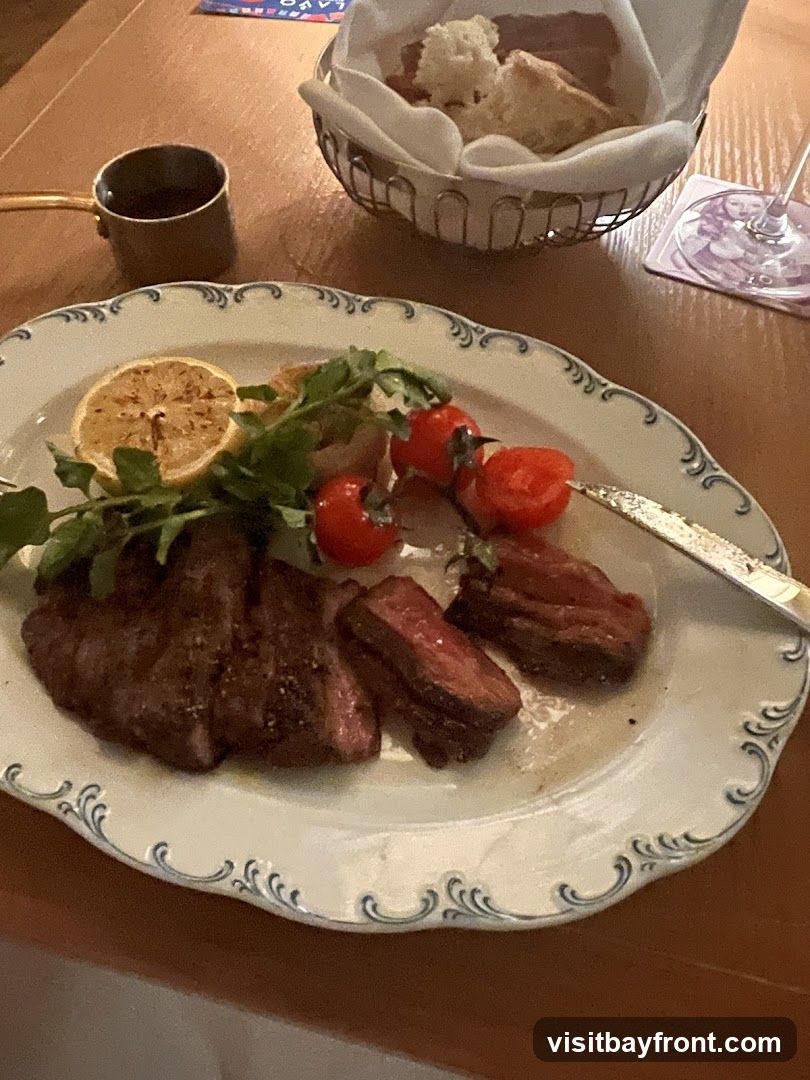 A plate of sliced steak is served with a lemon wedge, cherry tomatoes, and greens. A small metal pitcher of sauce and a basket of bread are also on the table.
