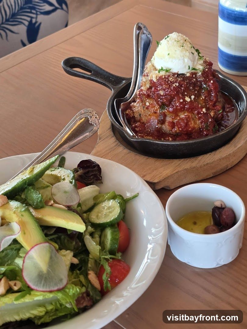 A large meatball covered in tomato sauce and cheese is served in a small cast iron skillet. Next to it is a salad with lettuce, avocado, cucumber, tomatoes, and radishes. A small dish of olives and dressing is also visible.